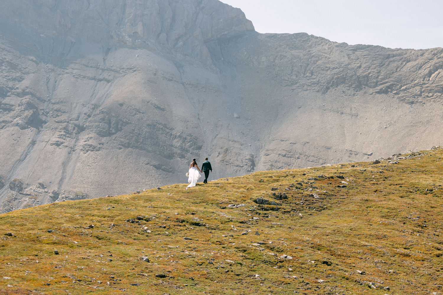 A couple holds hands while walking across a grassy hill with a rugged mountain backdrop.