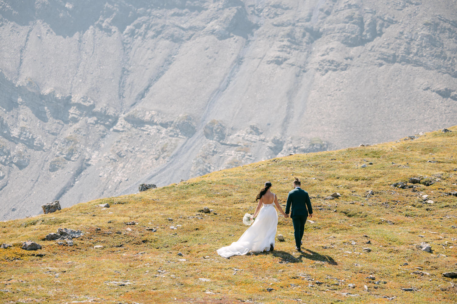 A couple walks hand in hand on a grassy hillside, surrounded by majestic mountains in the background.