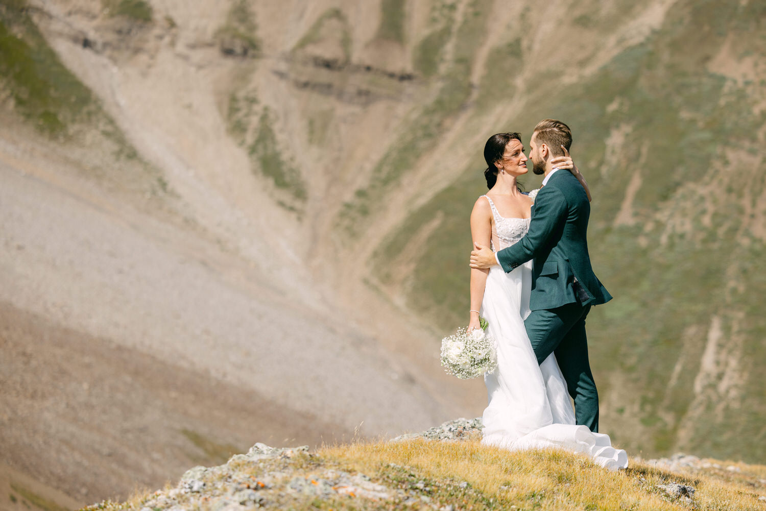 A bride and groom share a romantic moment on a mountainside, surrounded by stunning natural scenery.