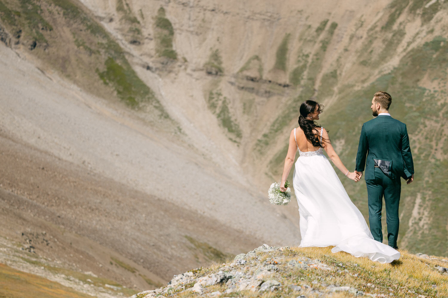 A couple holding hands on a grassy ledge, with mountains in the background, celebrating their wedding day. The bride holds a bouquet of flowers while wearing a flowing white dress, and the groom is dressed in a stylish dark suit.