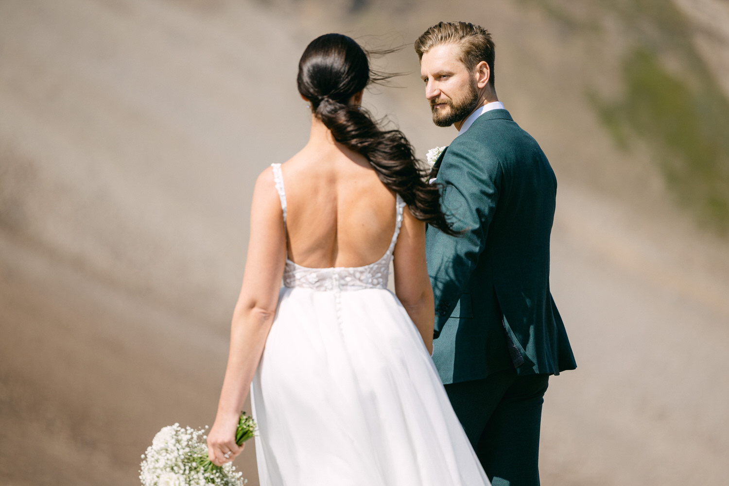 Romantic Outdoor Wedding Moment::A bride in a flowing white dress stands with her back to the camera, holding a bouquet, while a groom in a teal suit gazes at her, set against a natural landscape.