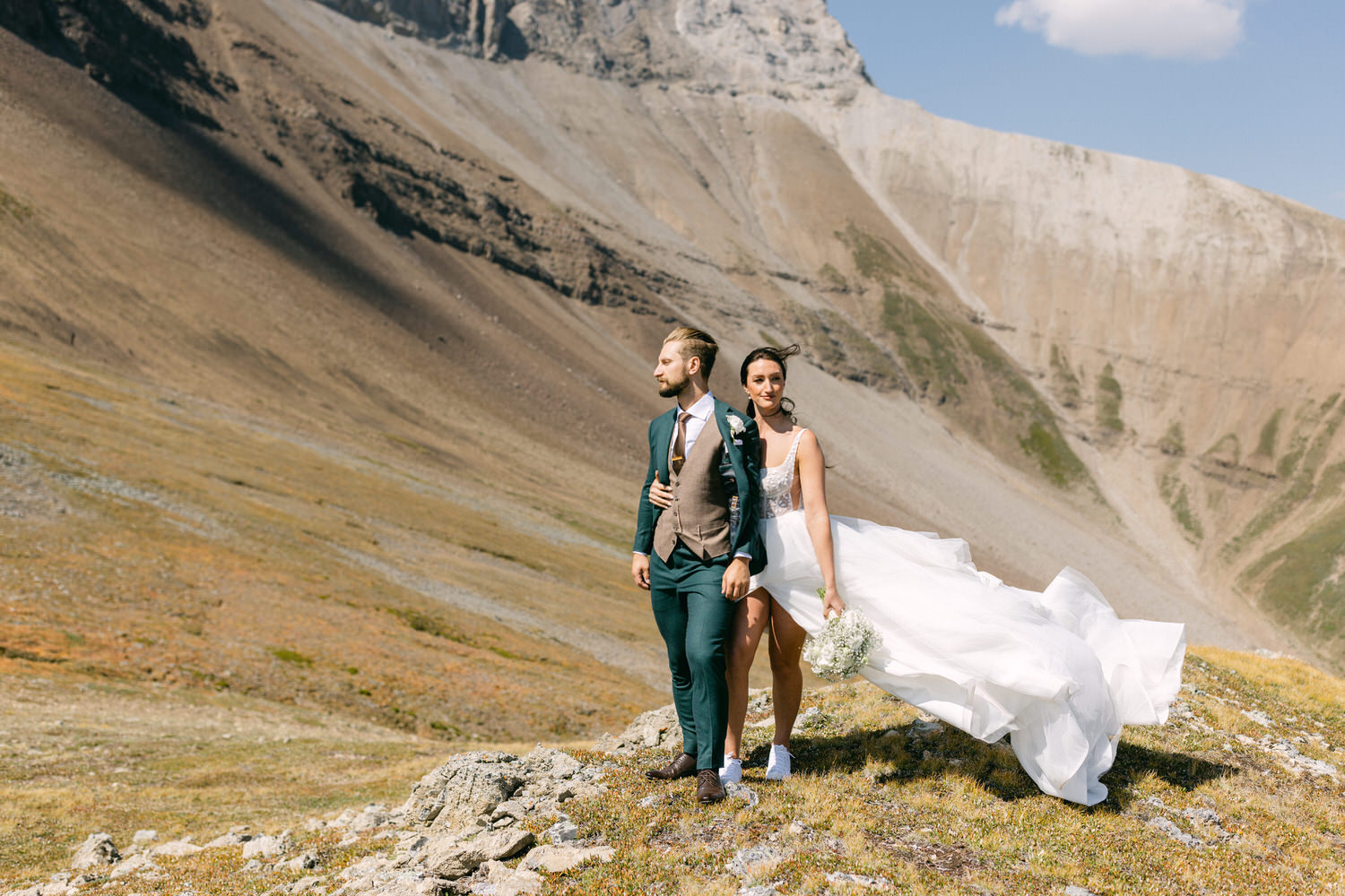 A couple in wedding attire stands on a grassy mountain slope, with a stunning backdrop of rocky hills and clear blue skies. The groom wears a teal suit with a brown vest, while the bride, holding a bouquet, has a flowing white gown and white sneakers.