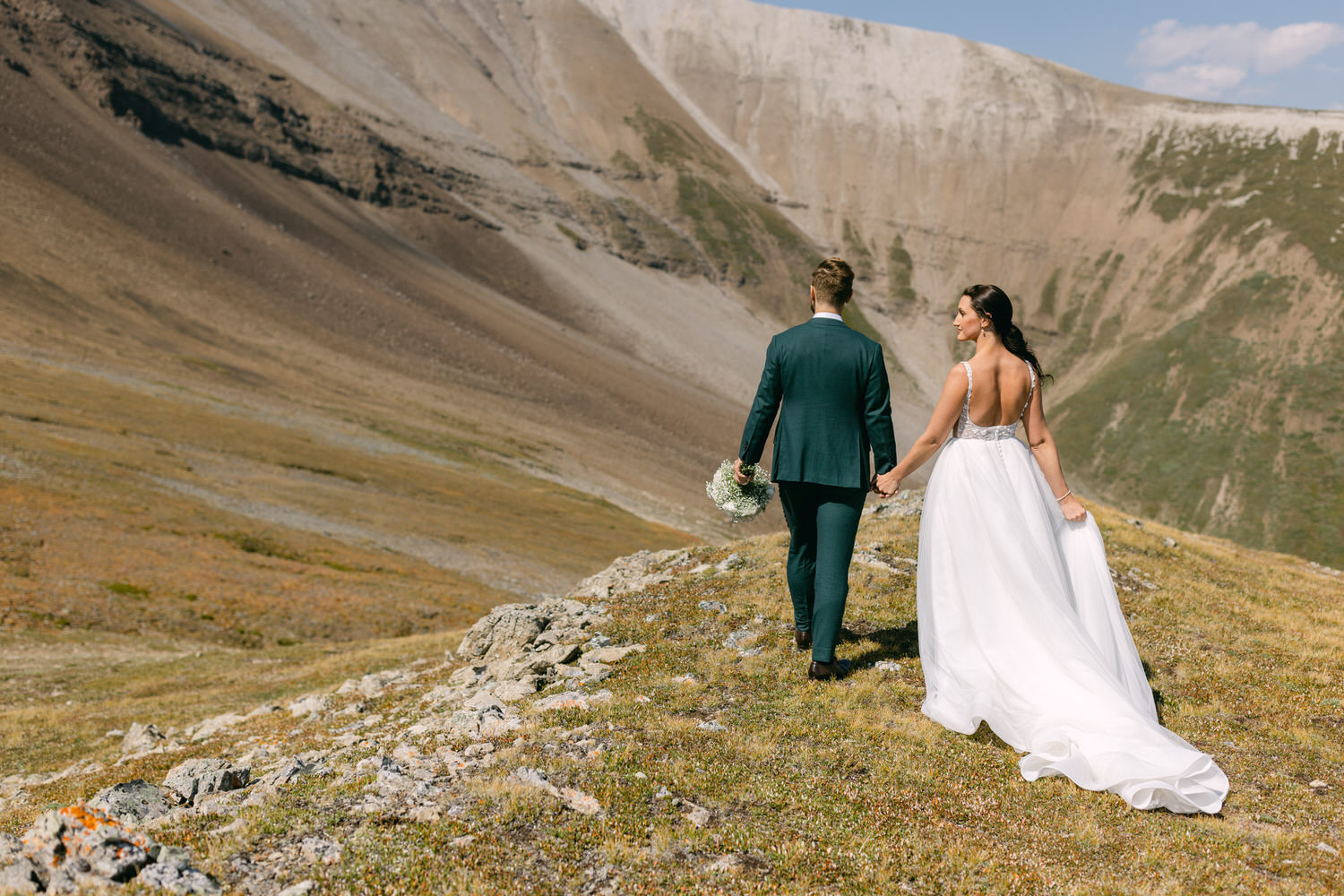 A couple in wedding attire walking hand in hand through a scenic mountainous landscape under a clear blue sky.