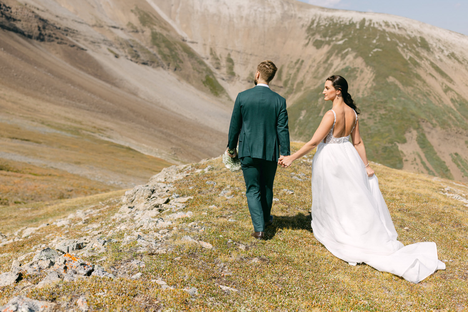 A bride and groom holding hands, walking along a rocky path in a mountainous landscape, wearing elegant wedding attire.