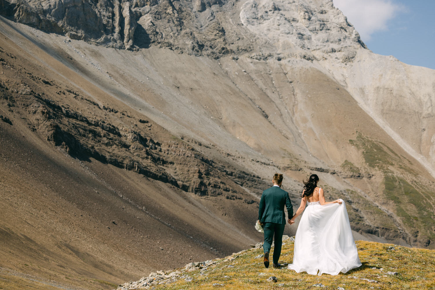 A couple walks hand-in-hand on a grassy slope with mountains in the background, showcasing a beautiful wedding scene.