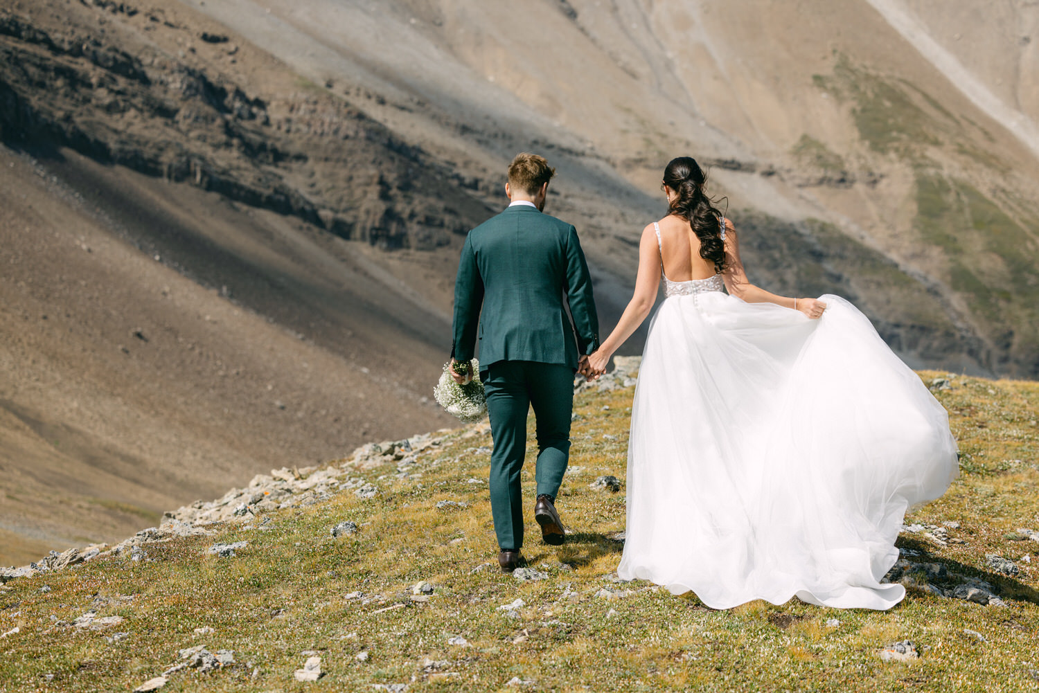 A couple in wedding attire walking hand in hand through a mountainous landscape, with the bride's flowing dress and bouquet adding a touch of elegance to the natural scenery.