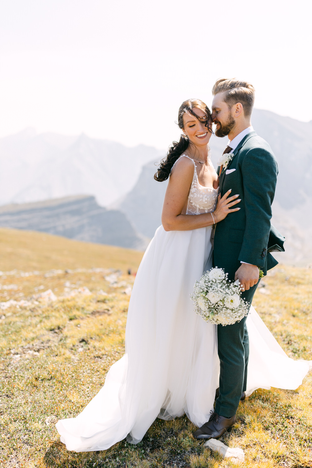 A joyful couple embraces in their wedding attire on a scenic mountaintop, surrounded by beautiful landscapes.