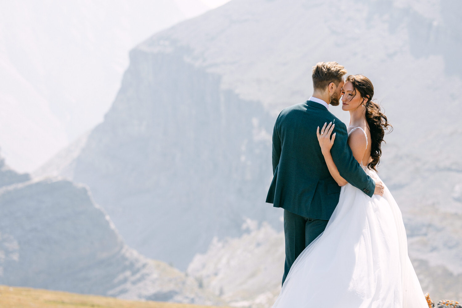 A couple in formal wedding attire shares a tender moment against a scenic mountain backdrop, radiating love and intimacy.