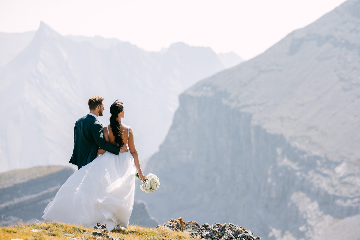 A couple in formal attire embraces while overlooking a stunning mountain landscape, capturing a moment of love and celebration.