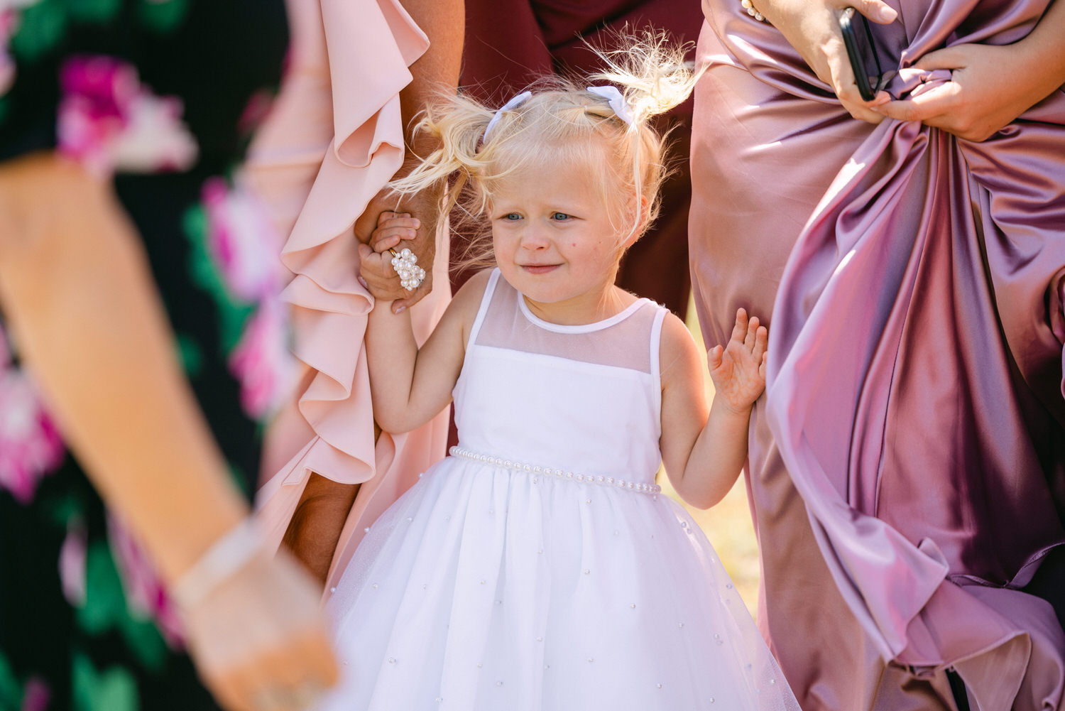 A young girl in a white dress with a pearl belt and blue hair bows, holding hands with adults in elegant dresses.