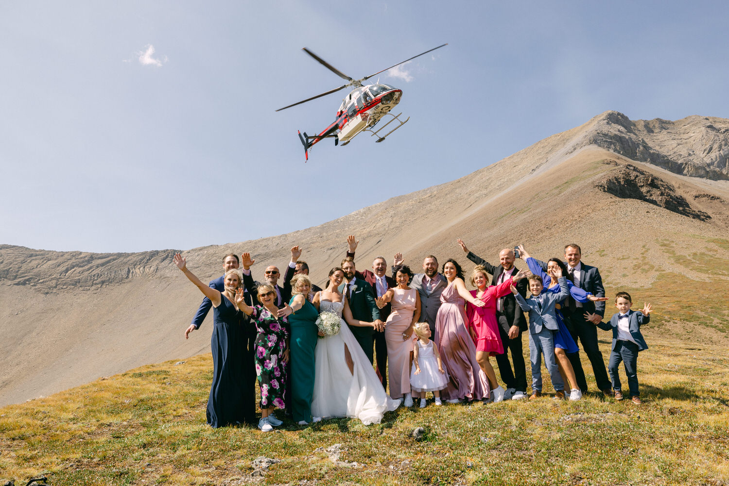 A joyful wedding party poses for a photo in the mountains, with a helicopter flying overhead, surrounded by stunning natural scenery.