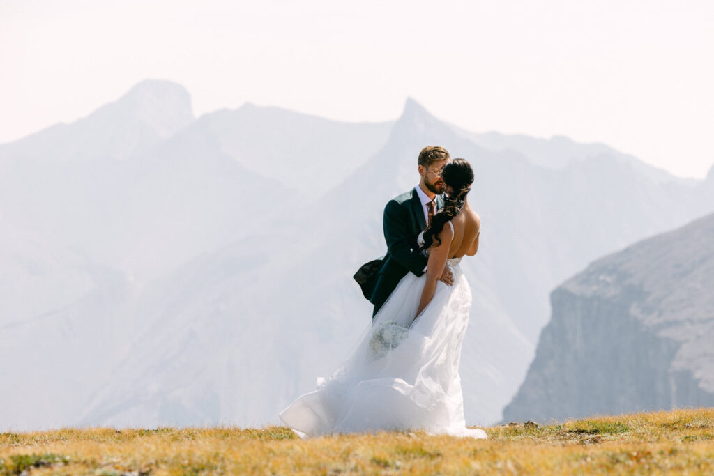 A couple shares a romantic kiss in a scenic mountain landscape, the bride in a flowing white gown and the groom in a suit, surrounded by majestic peaks.