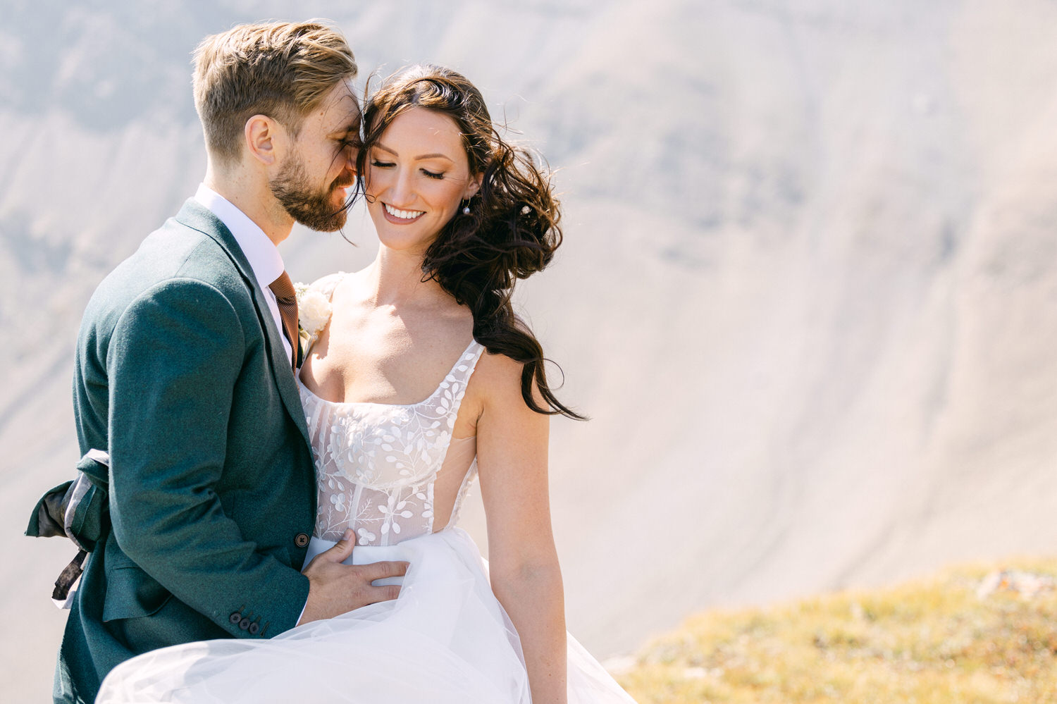 A joyful couple embraces in an outdoor setting, featuring a stunning bride in a floral dress and a groom in a stylish suit, with a beautiful mountain backdrop.