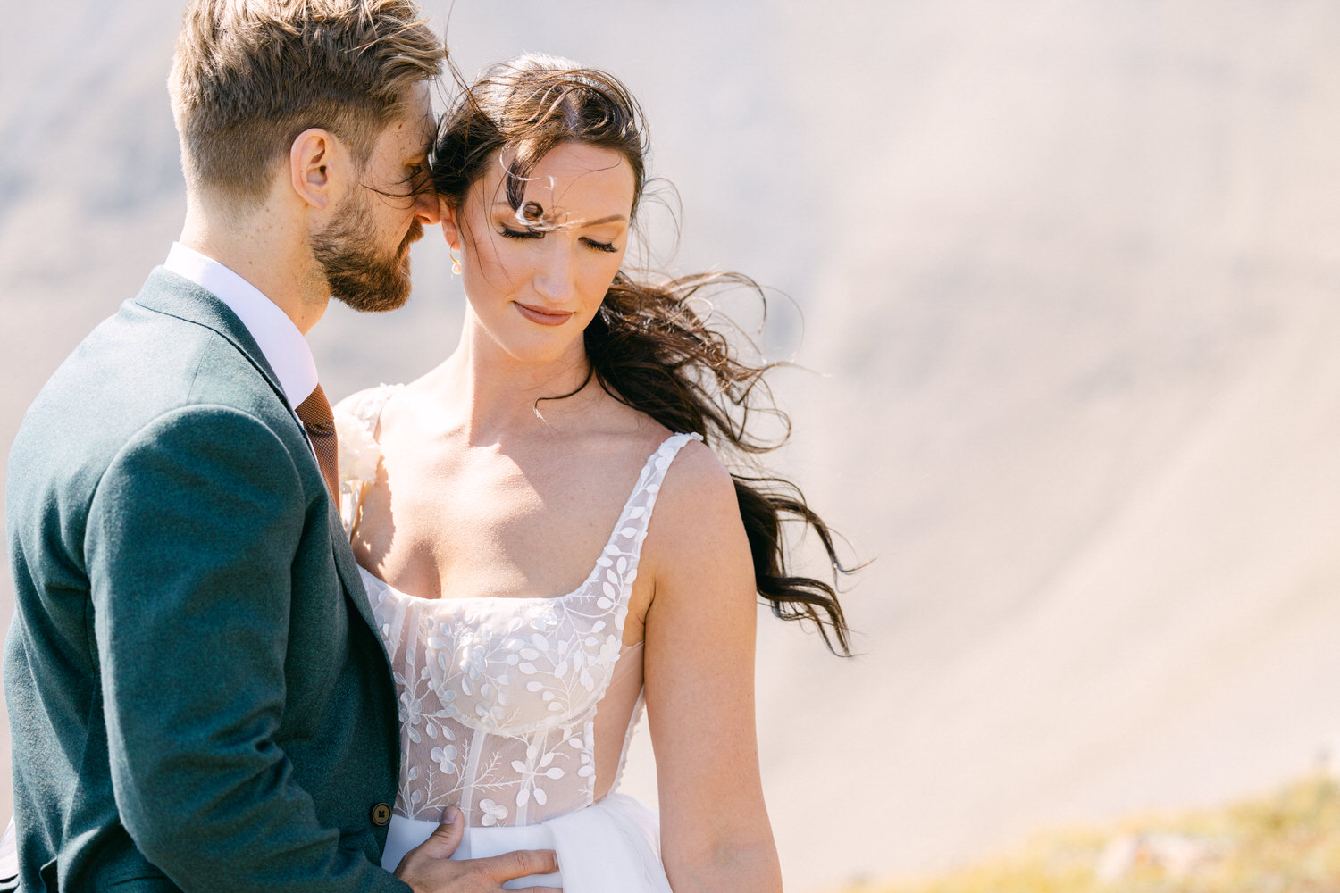 A couple in wedding attire shares an intimate moment, surrounded by a beautiful outdoor setting with flowing hair and gentle expressions.