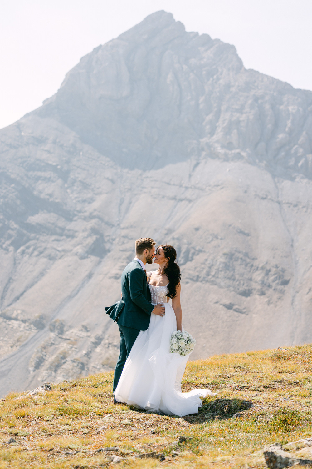 A couple embracing in a romantic kiss against a stunning mountain backdrop, showcasing love and nature on their special day.