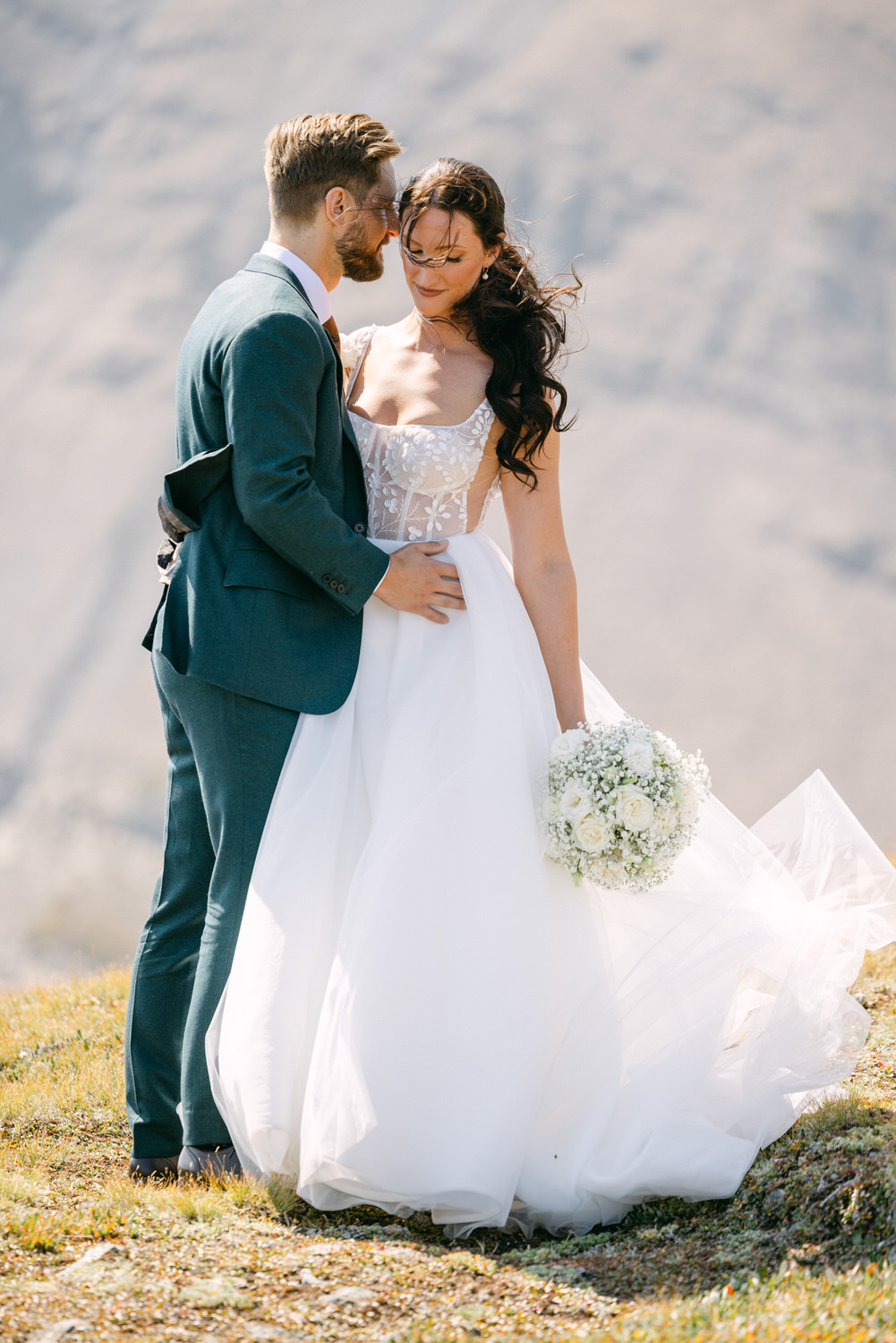 A bride and groom embrace in a picturesque outdoor setting, with the bride holding a bouquet of white flowers and wearing an elegant white wedding dress.