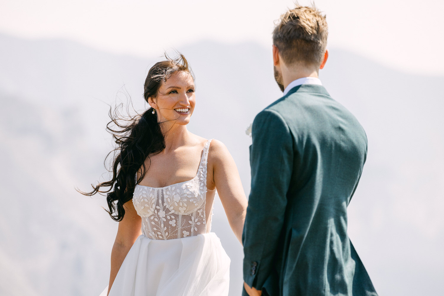 A bride smiles joyfully at her groom during an outdoor wedding ceremony, with windswept hair and a scenic mountain backdrop.