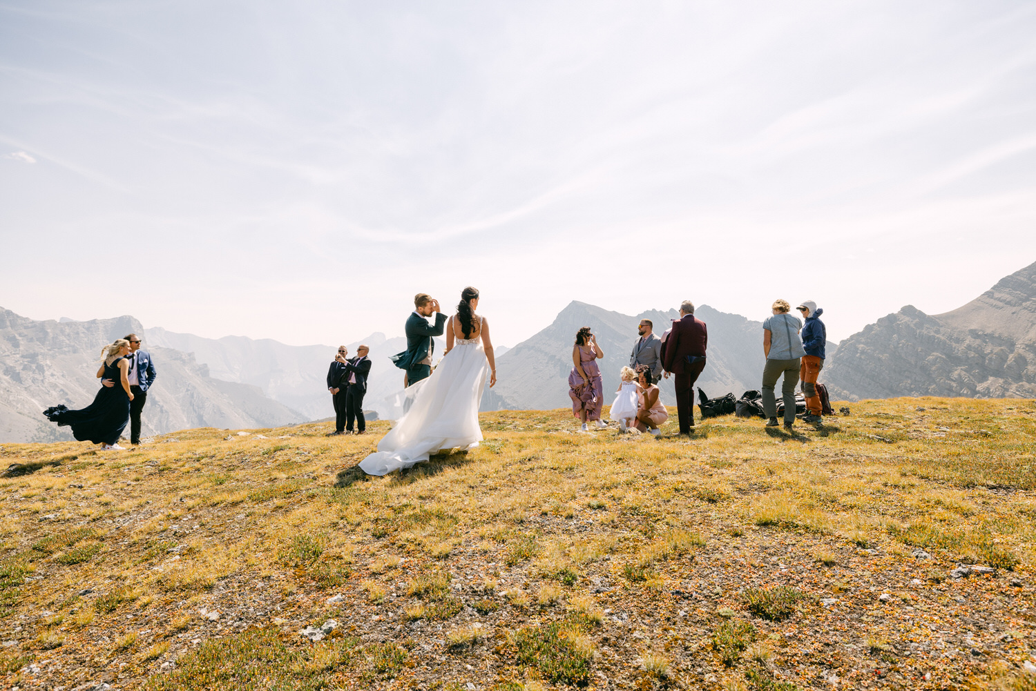 A picturesque mountain setting featuring wedding guests in formal attire, with two couples enjoying moments together amidst a stunning natural backdrop.