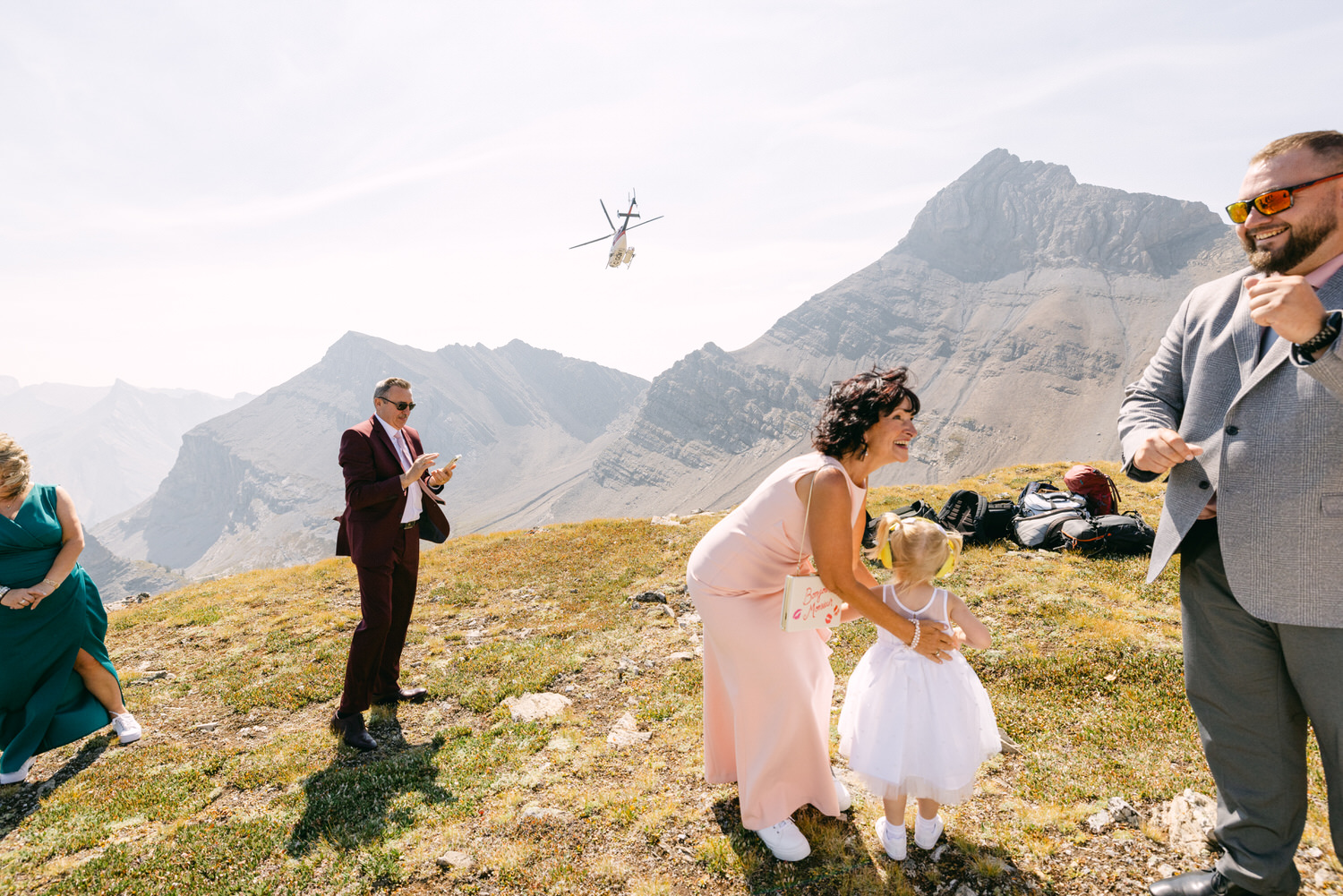 A joyful scene of guests in elegant attire celebrating on a mountain as a helicopter flies overhead, surrounded by stunning mountain scenery.