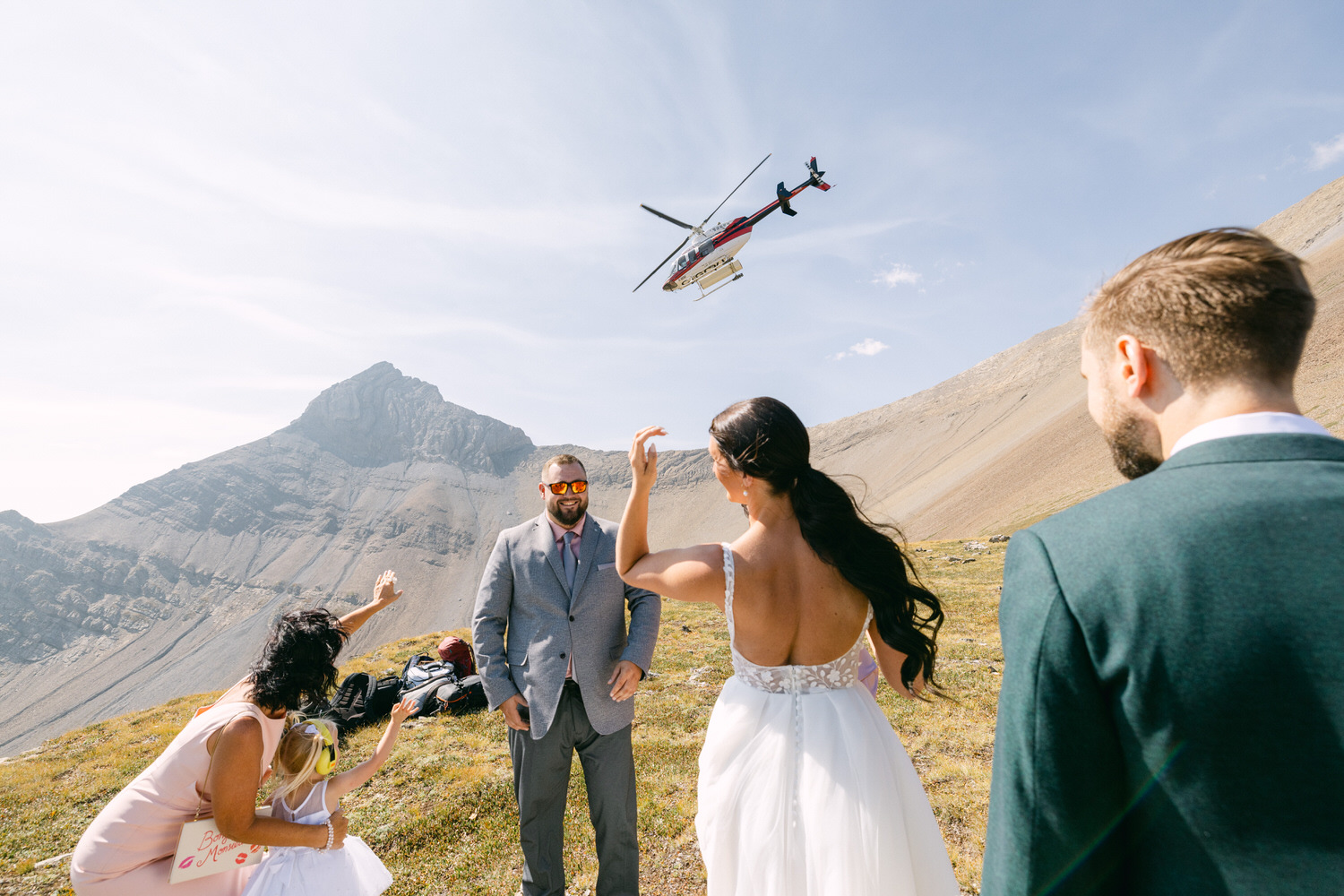 A bride and groom greet guests as a helicopter flies overhead in a picturesque mountain setting.