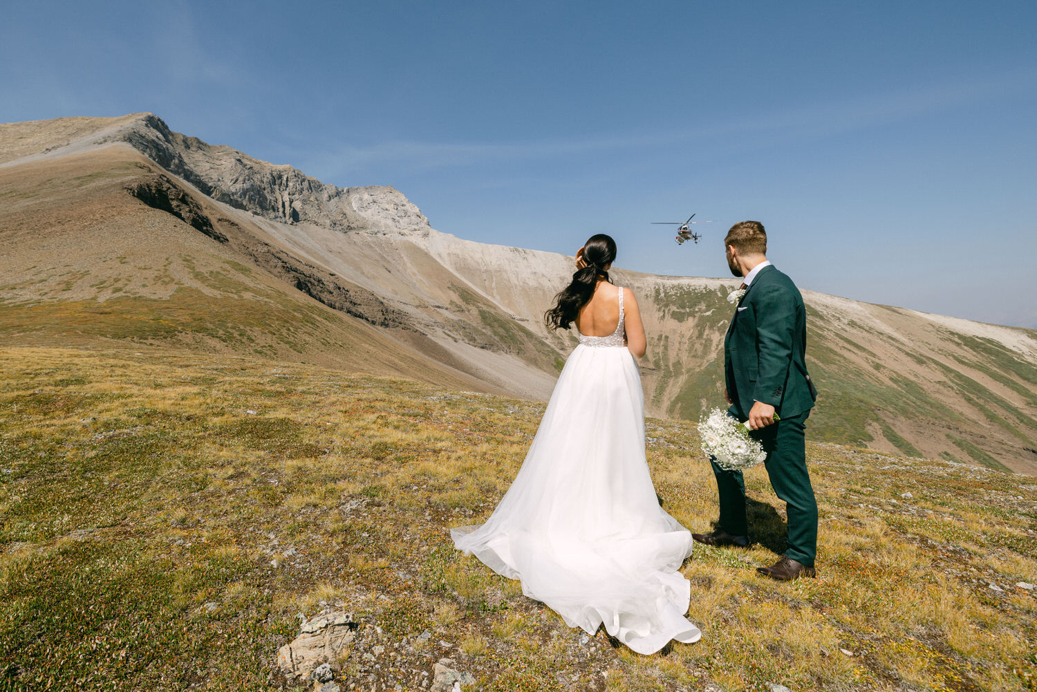  A bride and groom stand on a grassy hillside, overlooking a mountainous landscape, as a helicopter flies overhead.