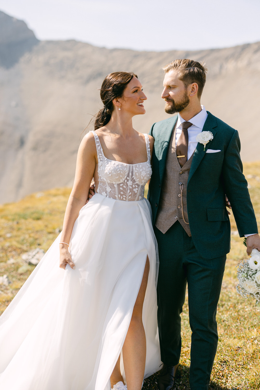 A smiling couple dressed in wedding attire stands together in a scenic outdoor setting, surrounded by mountains and greenery.