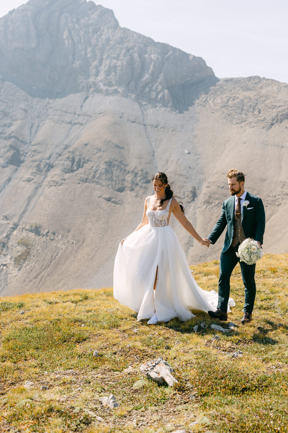 A couple holds hands while walking in a scenic mountainous landscape; the bride in a flowing white gown and the groom in a tailored suit, surrounded by nature.