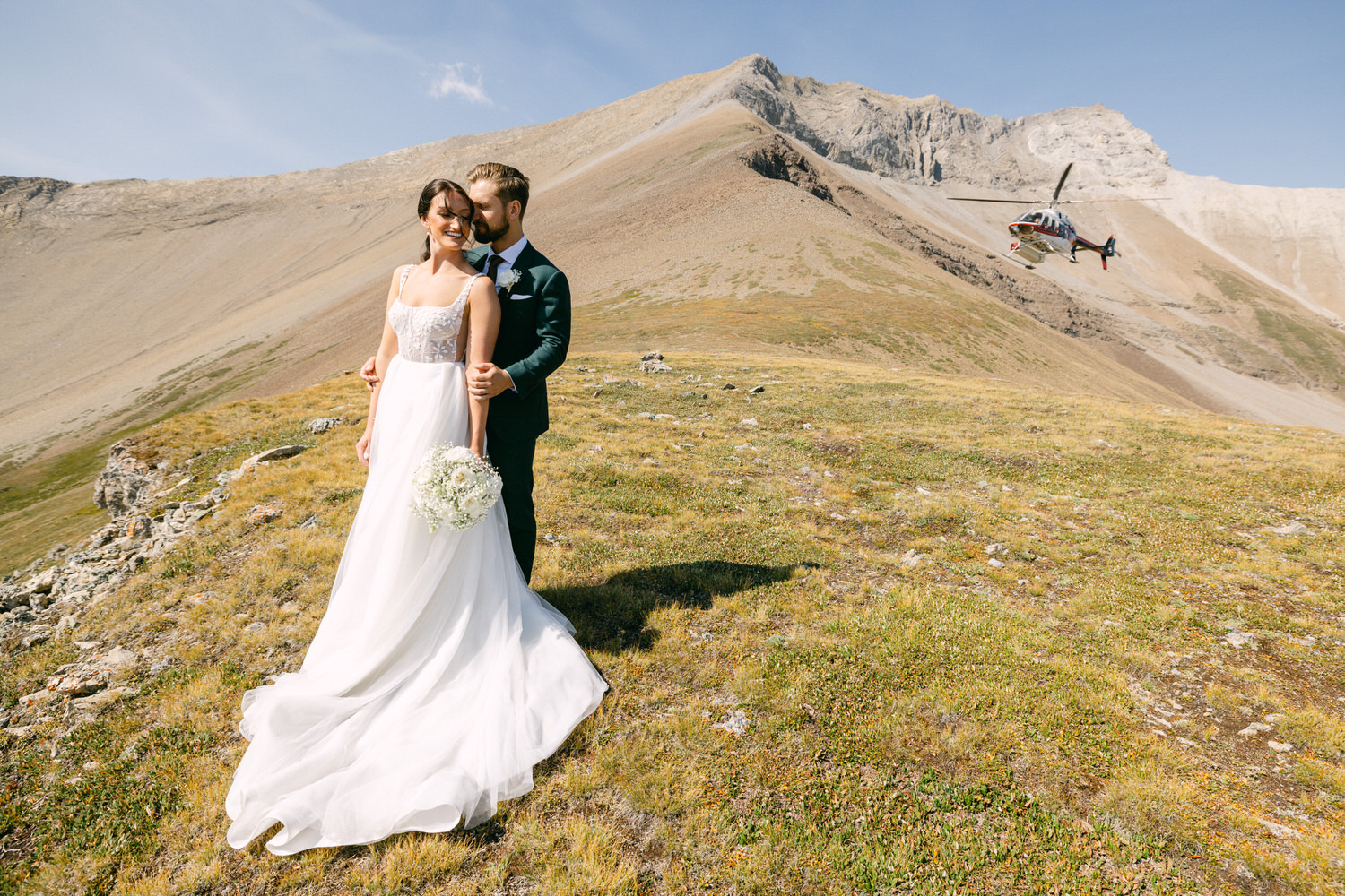 A couple embraces on a mountainside during their wedding, with a helicopter flying in the background under a bright blue sky.