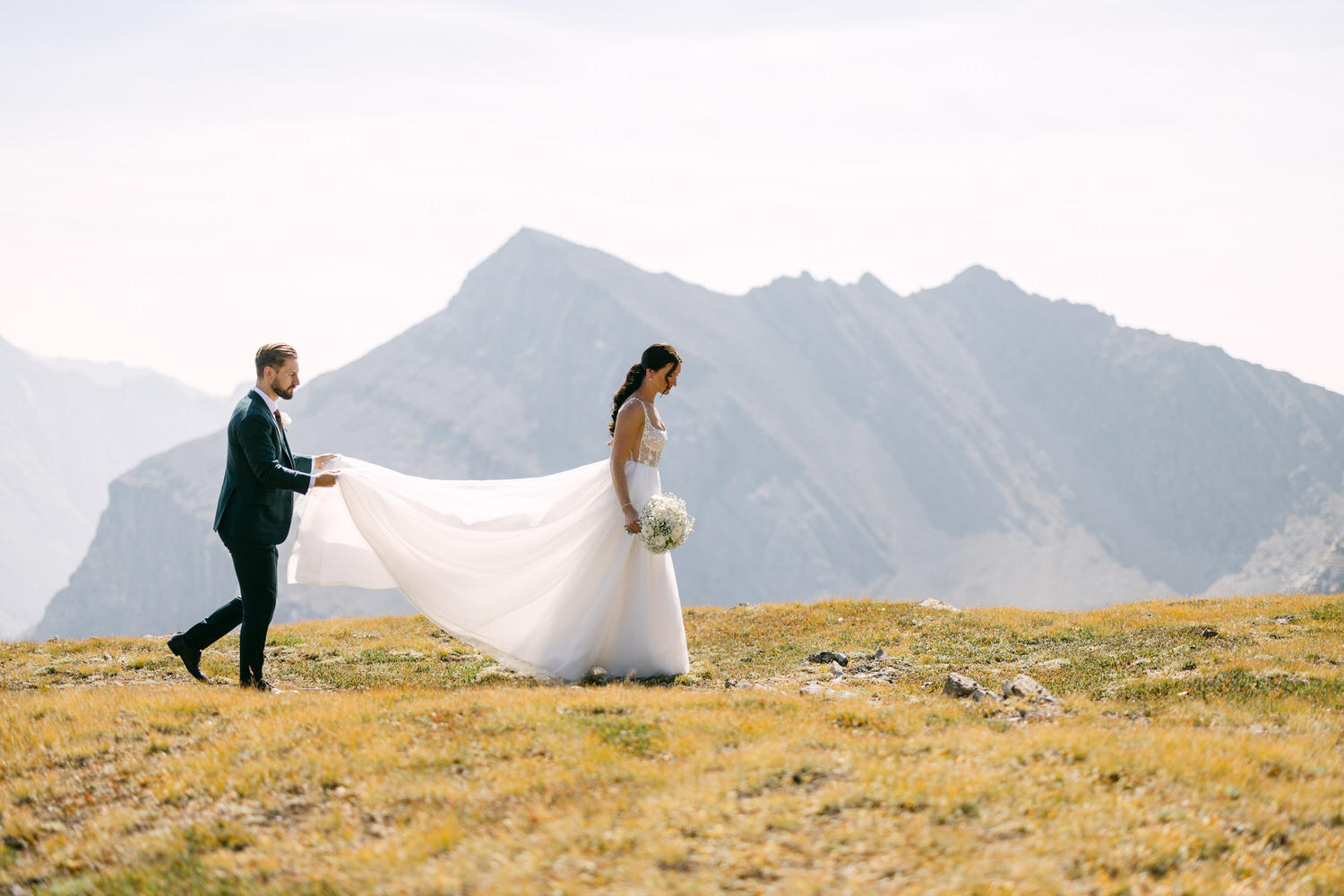 Mountain Bridal Portrait: A couple sharing a beautiful moment in a scenic mountain landscape during their wedding. The bride's elegant gown flows in the gentle breeze, while the groom supports her train as they walk.