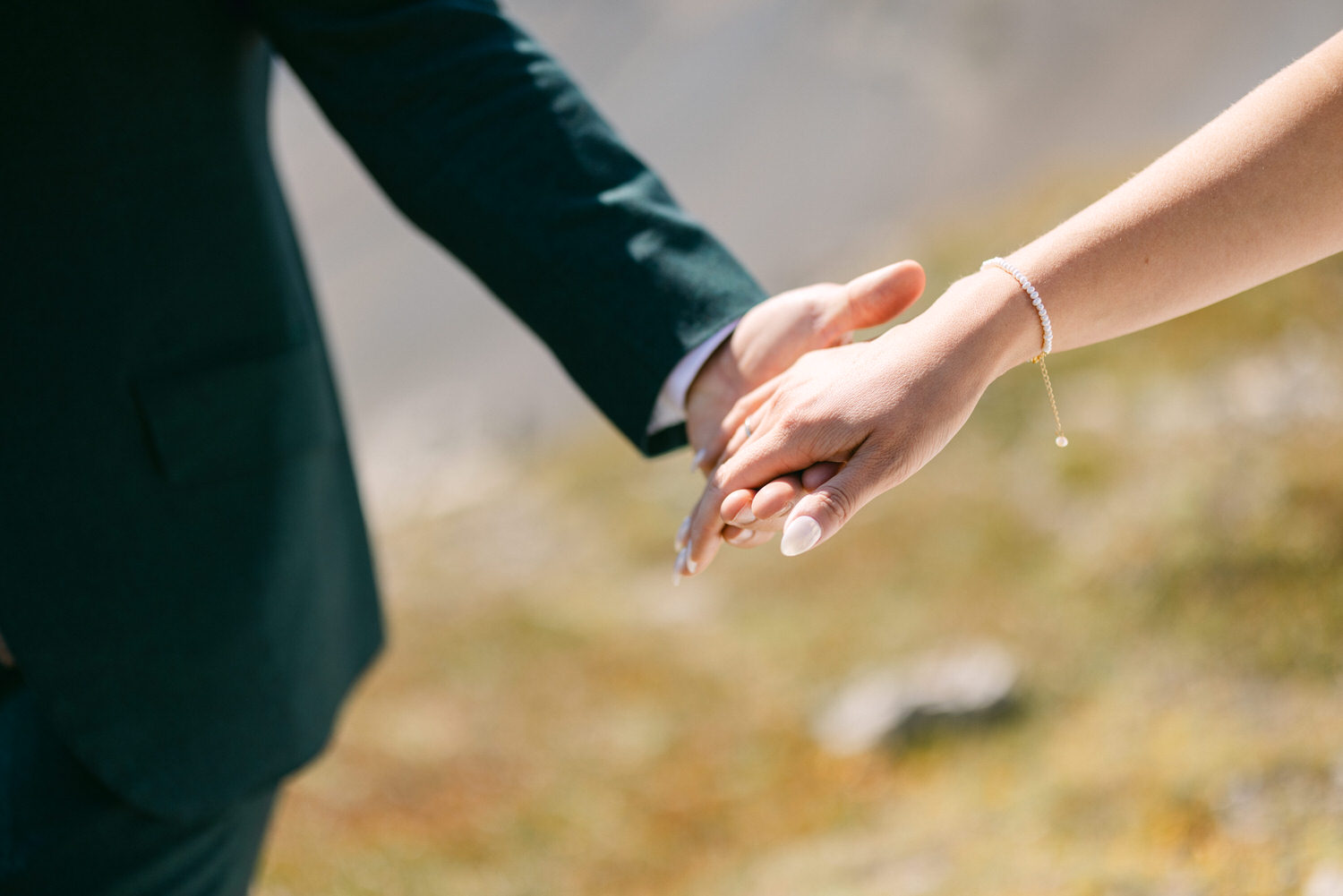 Close-up of two hands holding each other, one adorned with a bracelet, against a blurred outdoor backdrop.