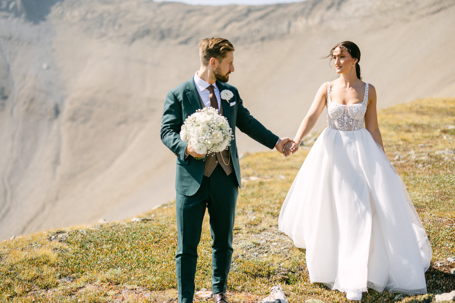 A bride and groom holding hands on a scenic mountainside, with the bride in a white gown and the groom in a dark suit, surrounded by nature.