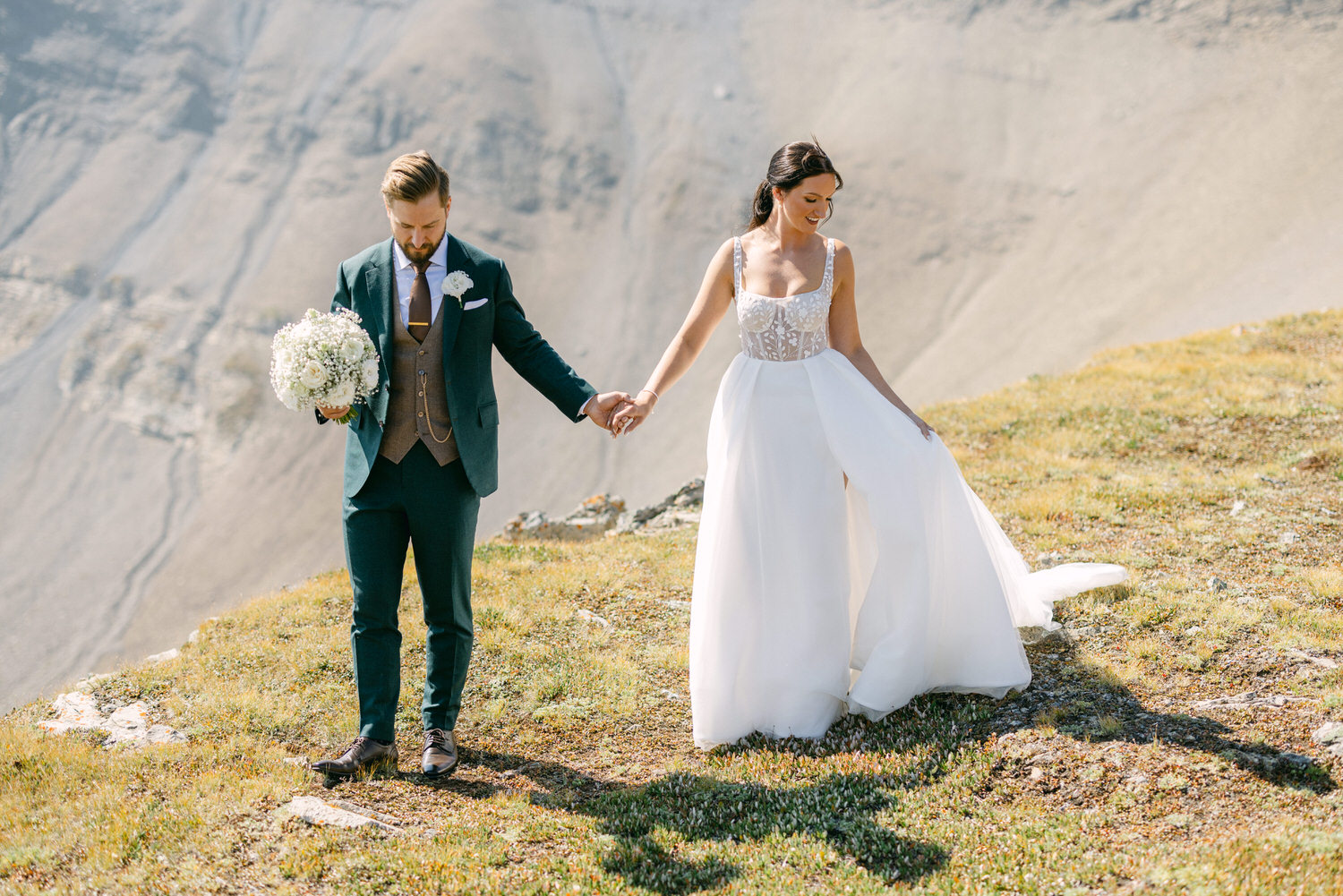 A couple holding hands on a mountain meadow, the bride in a flowing white gown and the groom in a stylish suit, surrounded by natural beauty.