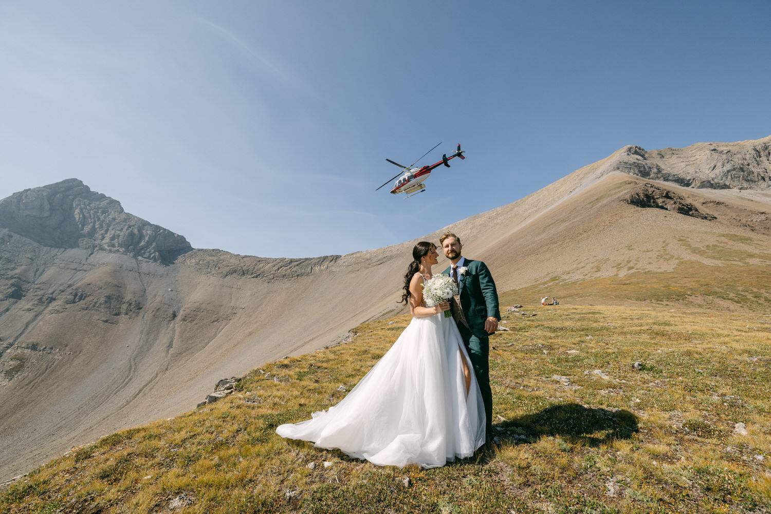 A couple embraces on a scenic mountain landscape as a helicopter flies overhead, capturing the essence of their unique wedding day.