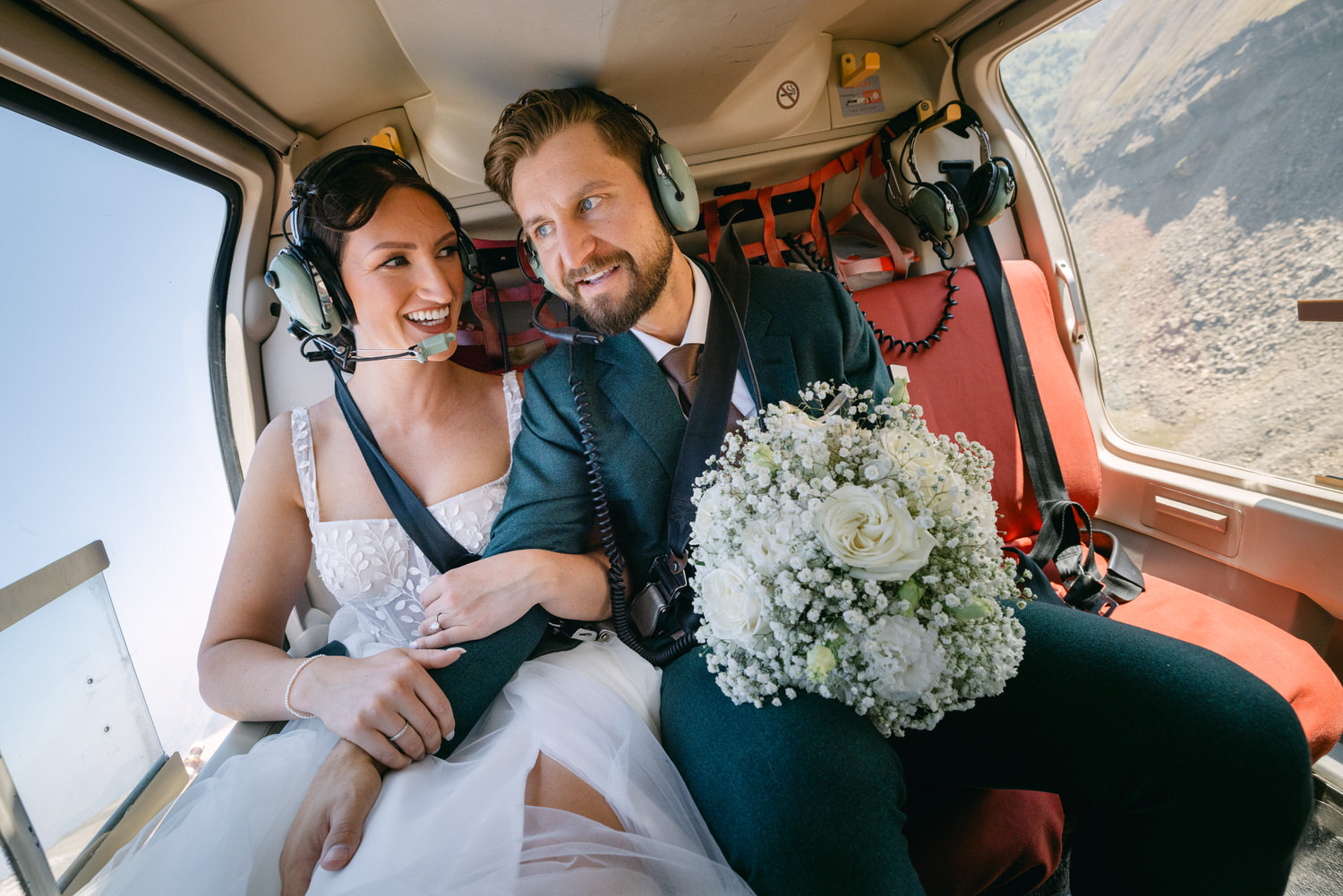 A joyful couple in wedding attire enjoying a helicopter ride, smiling at each other while holding a bouquet of flowers.