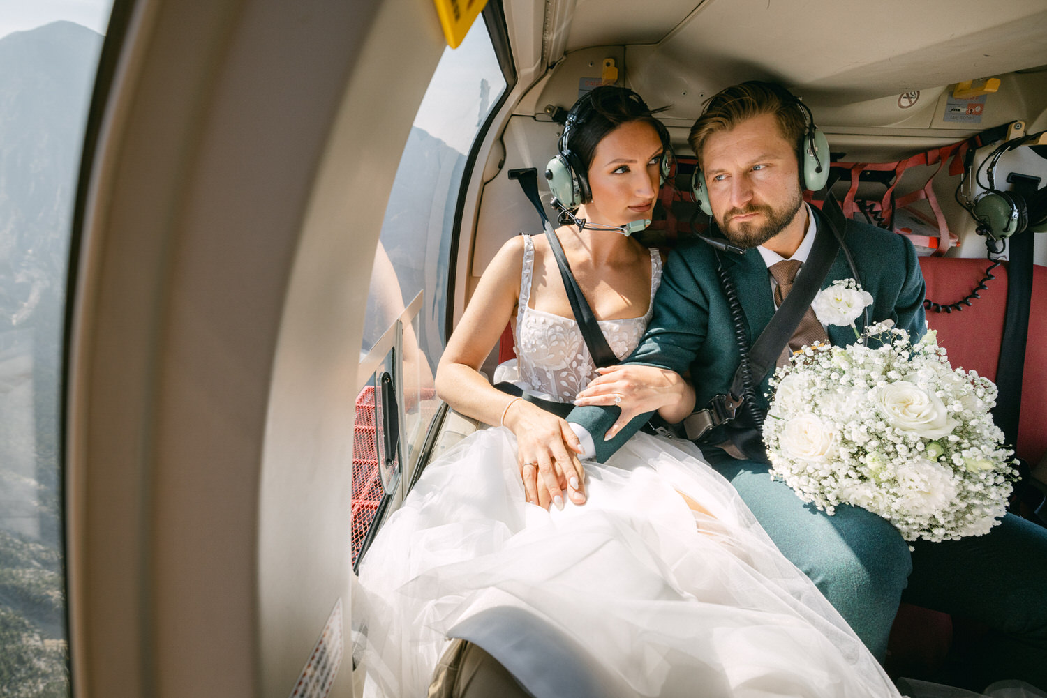 A bride and groom in a helicopter, wearing headsets, holding hands, and surrounded by stunning mountain views.