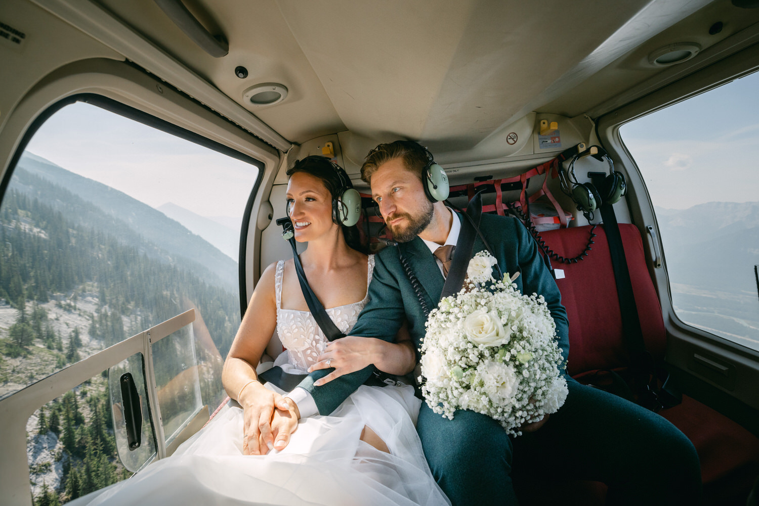 A happy couple, dressed in wedding attire, enjoying a scenic helicopter ride with a beautiful bouquet, surrounded by mountains.