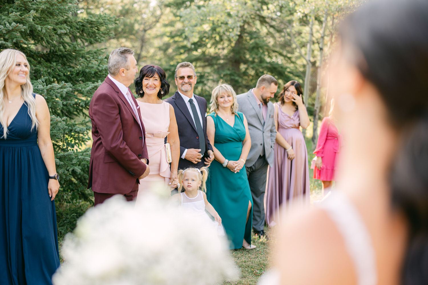 A group of elegantly dressed guests attending a wedding ceremony outdoors, surrounded by greenery, with a young girl in a white dress in the foreground.