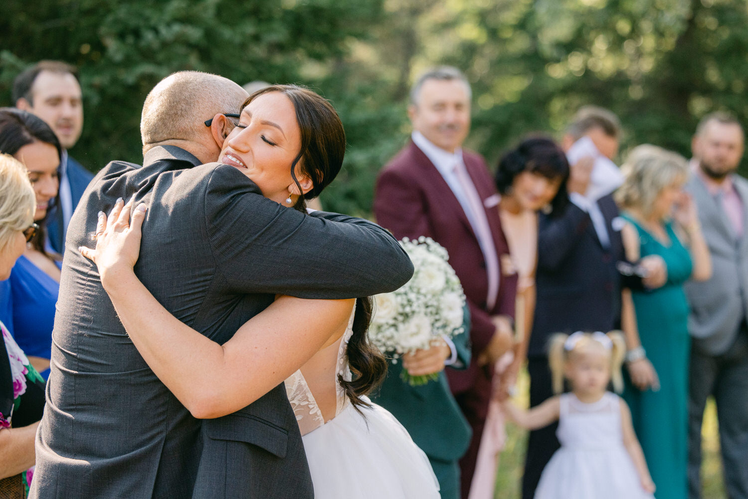 A bride hugs a man in a suit, surrounded by wedding guests, with a joyful expression in a beautiful outdoor setting.