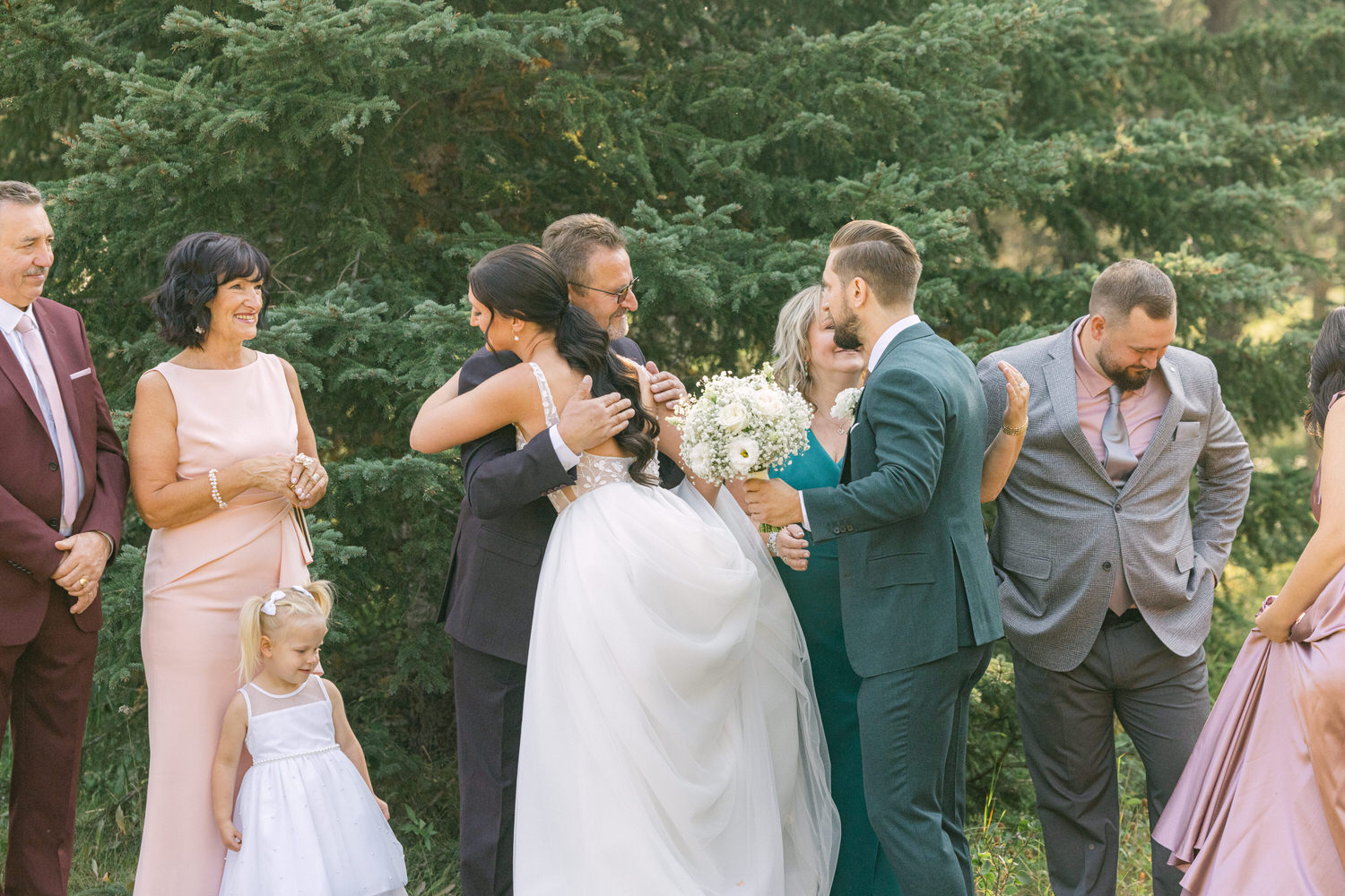 A bride hugs her father while surrounded by family members, celebrating a joyful wedding moment amidst lush greenery.