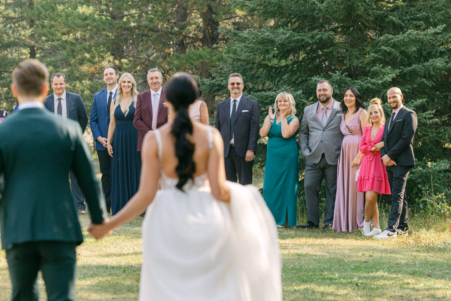 A bride and groom hold hands, facing a joyful gathering of family and friends in a lush, green outdoor setting.