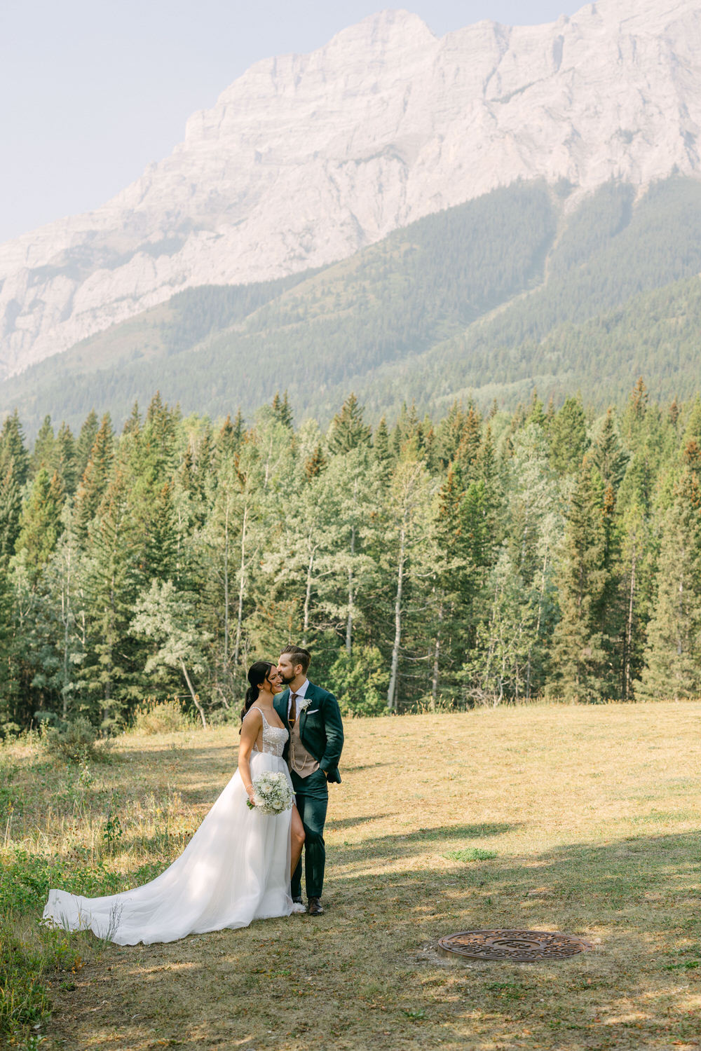 A bride and groom share a kiss in a serene outdoor setting surrounded by lush trees and majestic mountains.