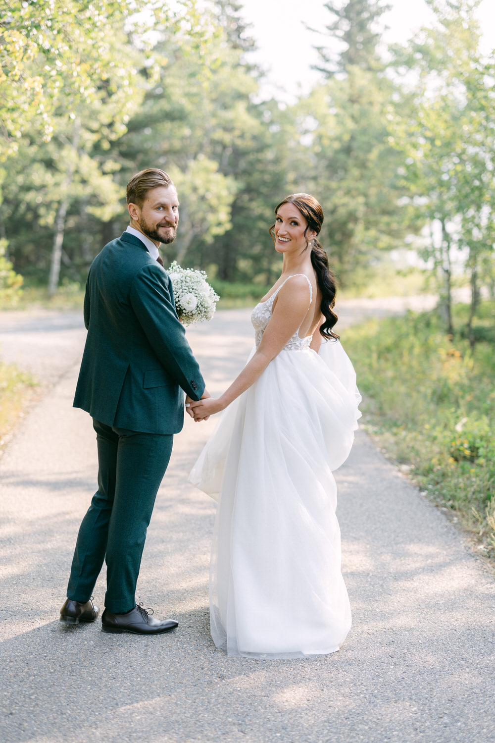 A couple holding hands while looking back at the camera, surrounded by lush greenery on a scenic path. The woman wears a flowing white wedding dress, and the man is in a dark suit, both smiling warmly.