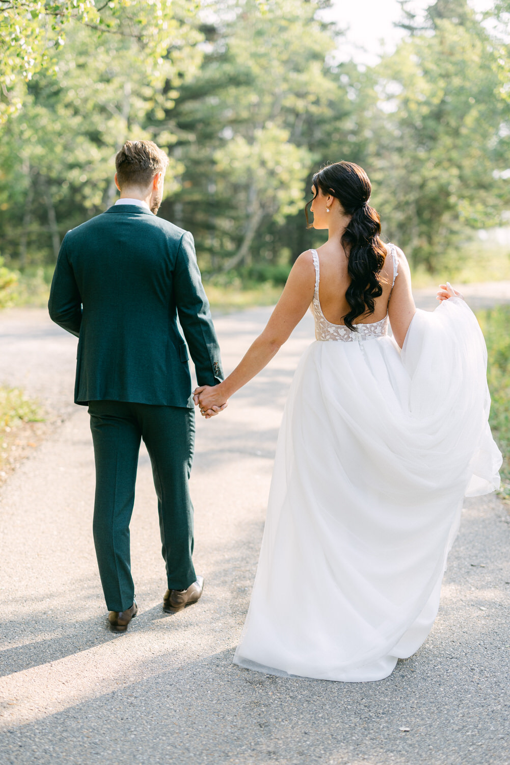 A bride in a flowing white gown and a groom in a dark suit stroll together along a scenic path surrounded by greenery.