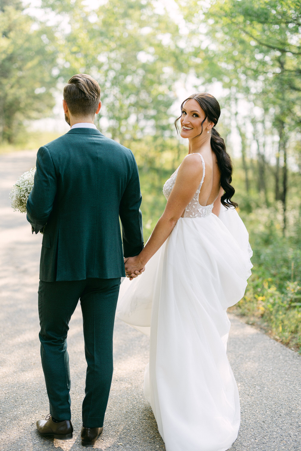 A happy couple holding hands on a scenic path, the bride in a flowing white gown and the groom in a dark suit, surrounded by greenery.