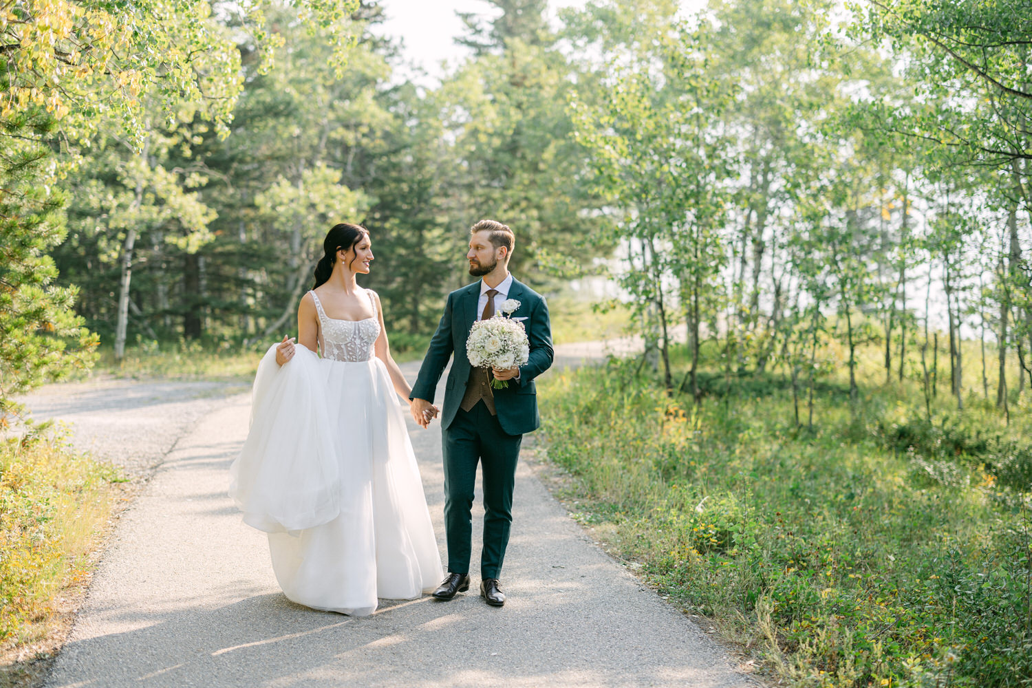 A smiling couple holding hands while walking along a tree-lined path, the bride in a flowing white gown and the groom in a smart suit, surrounded by greenery.