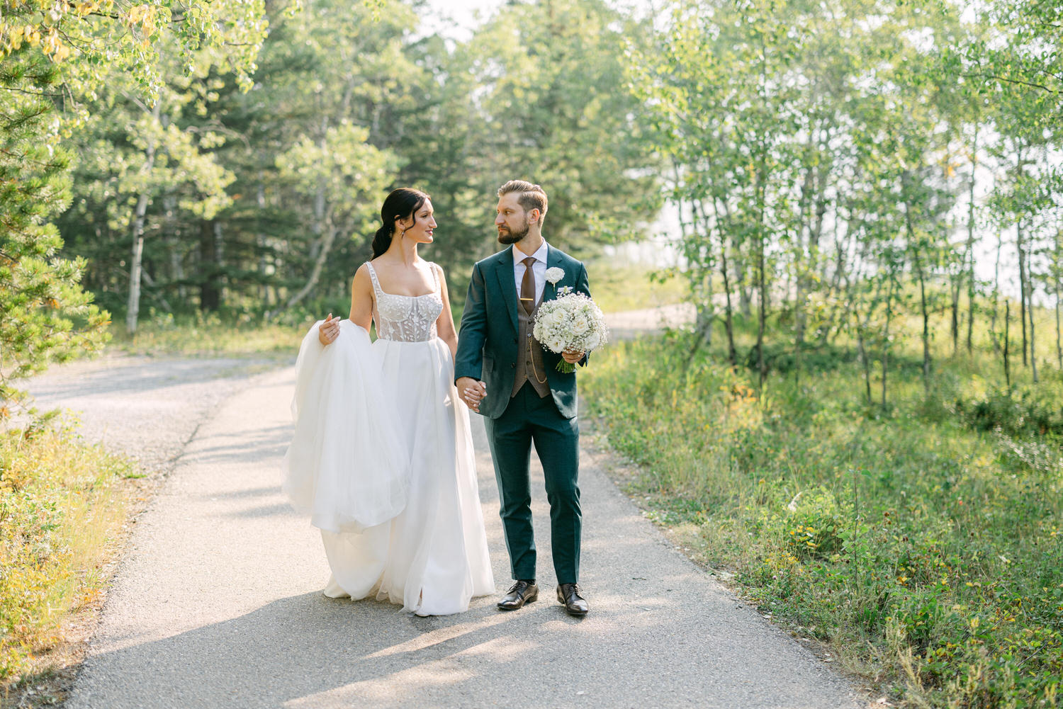 A bride in a flowing white gown and a groom in a navy suit stroll together along a tree-lined path, sharing a moment of joy in a natural setting.