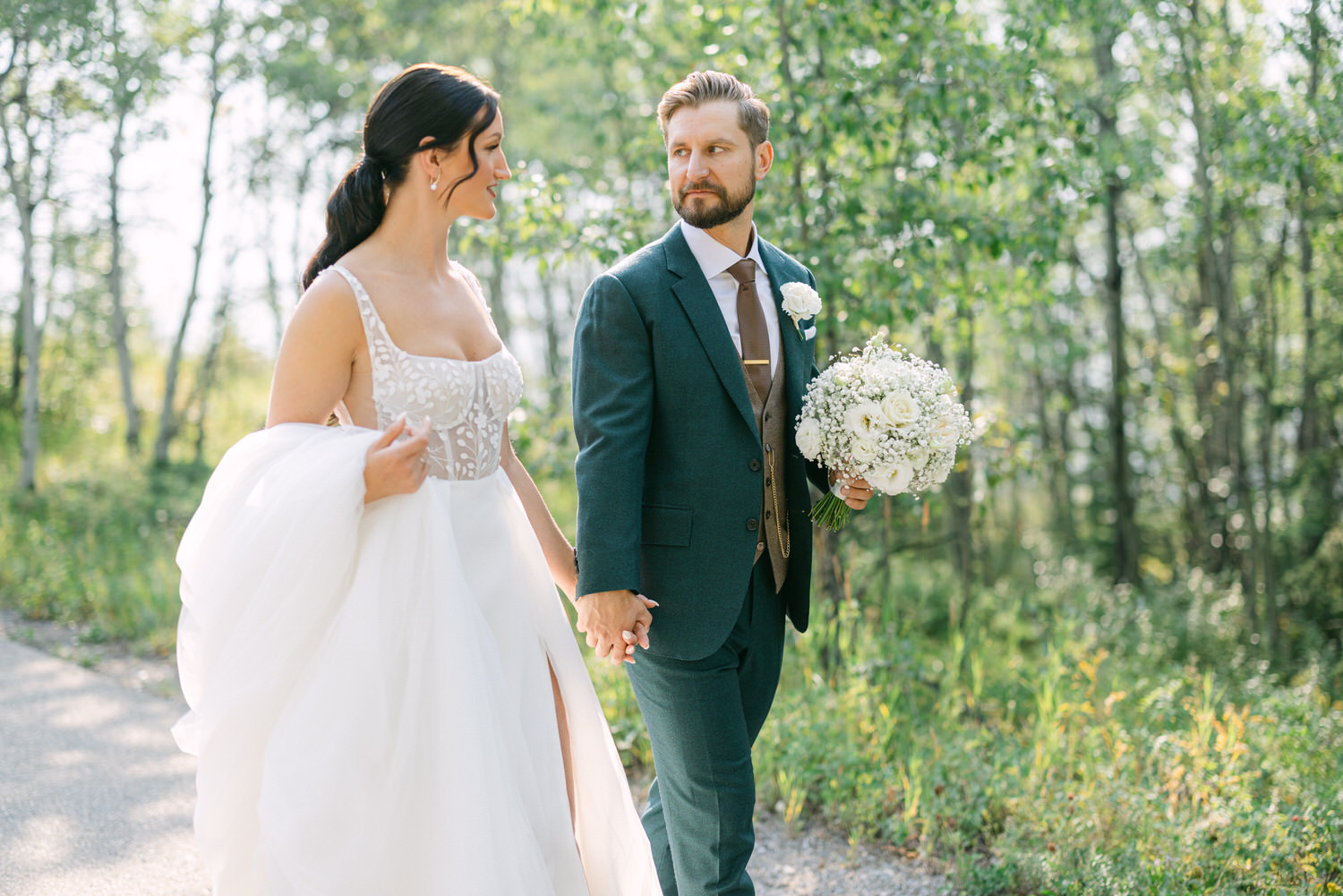 A bride in a stunning white gown and a groom in a dark suit stroll hand in hand through a lush green forest, both smiling and enjoying the moment.