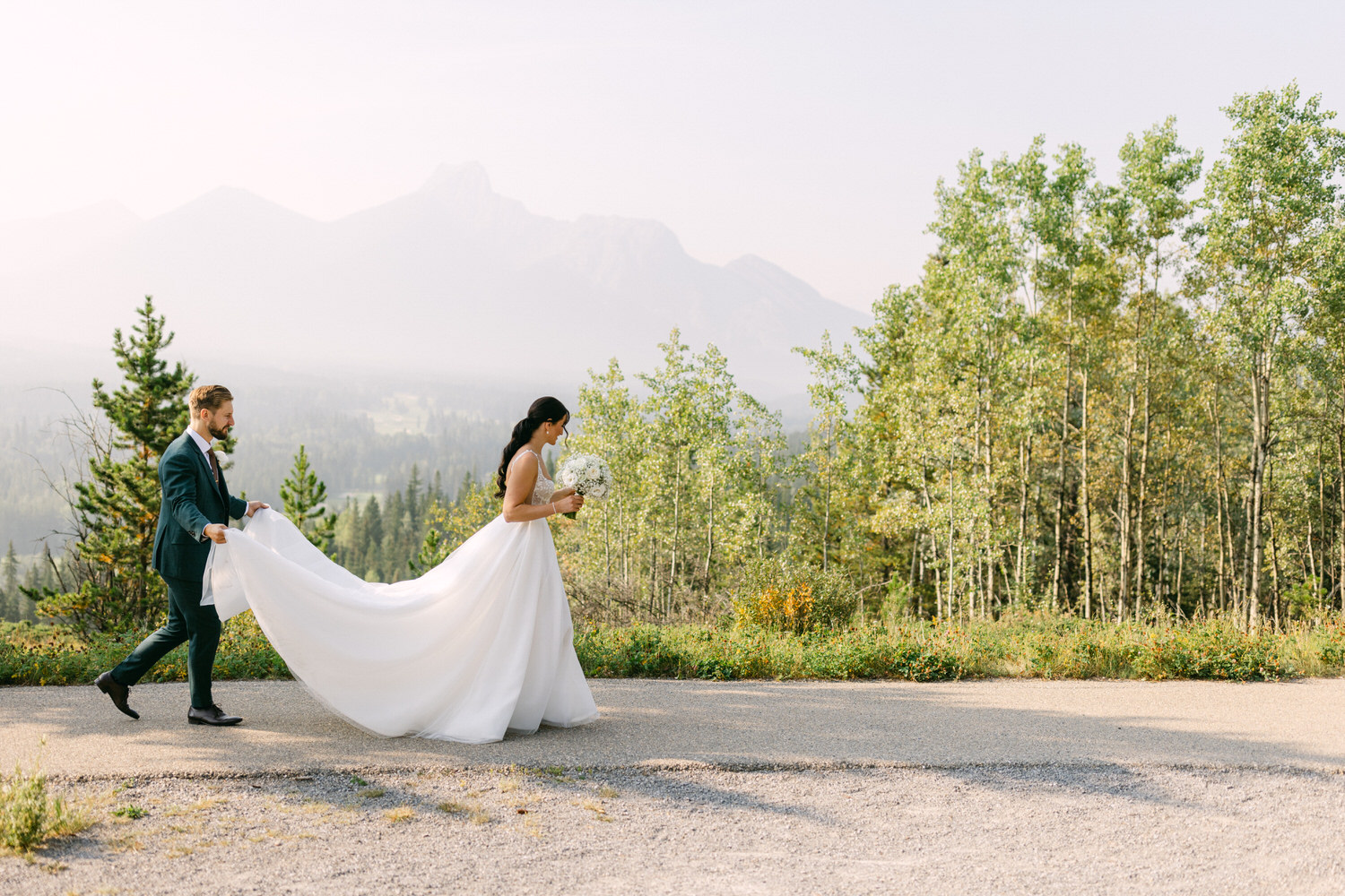 A bride in a flowing white gown walks on a path with her groom, who holds her train, amidst a scenic backdrop of mountains and greenery.