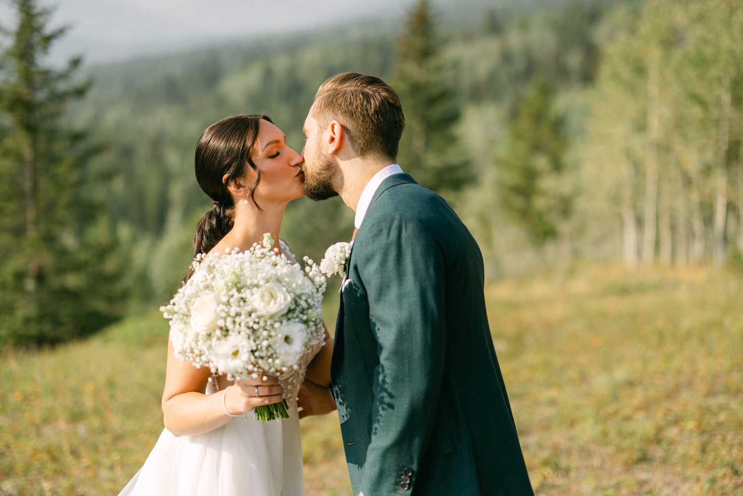 A bride and groom share a tender kiss amidst a natural, green landscape, with the bride holding a bouquet of white flowers.