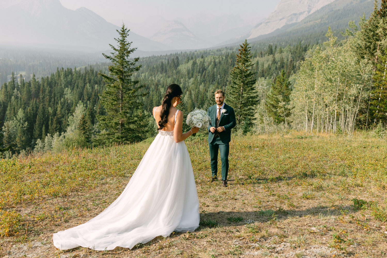 A bride in a flowing white gown holds a bouquet while walking towards her groom in a green suit, set against a breathtaking mountain and forest backdrop.